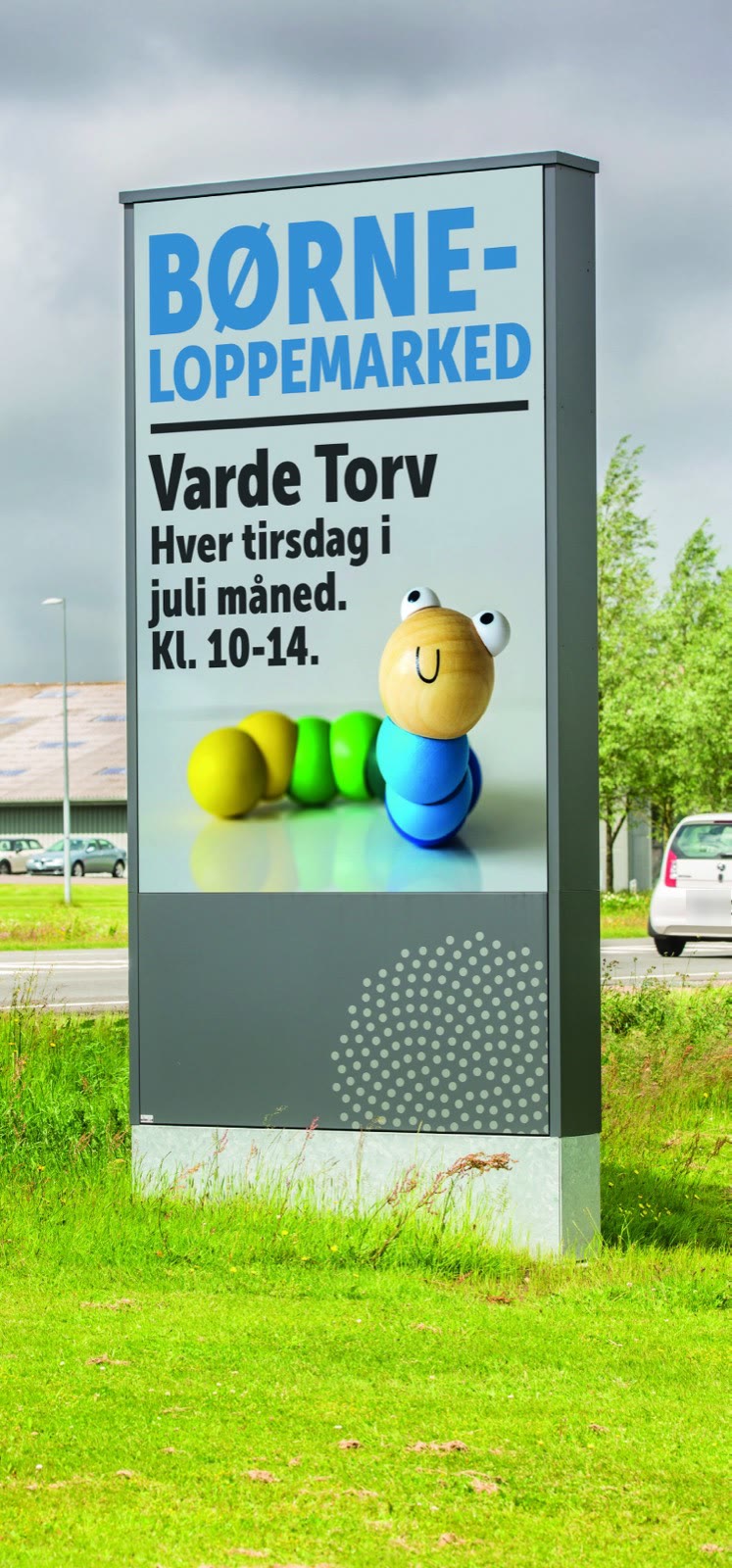 LED-pylon i græsset ved en vej. Skærmen viser en plakat, og under den er et gråt panel med prikmønster